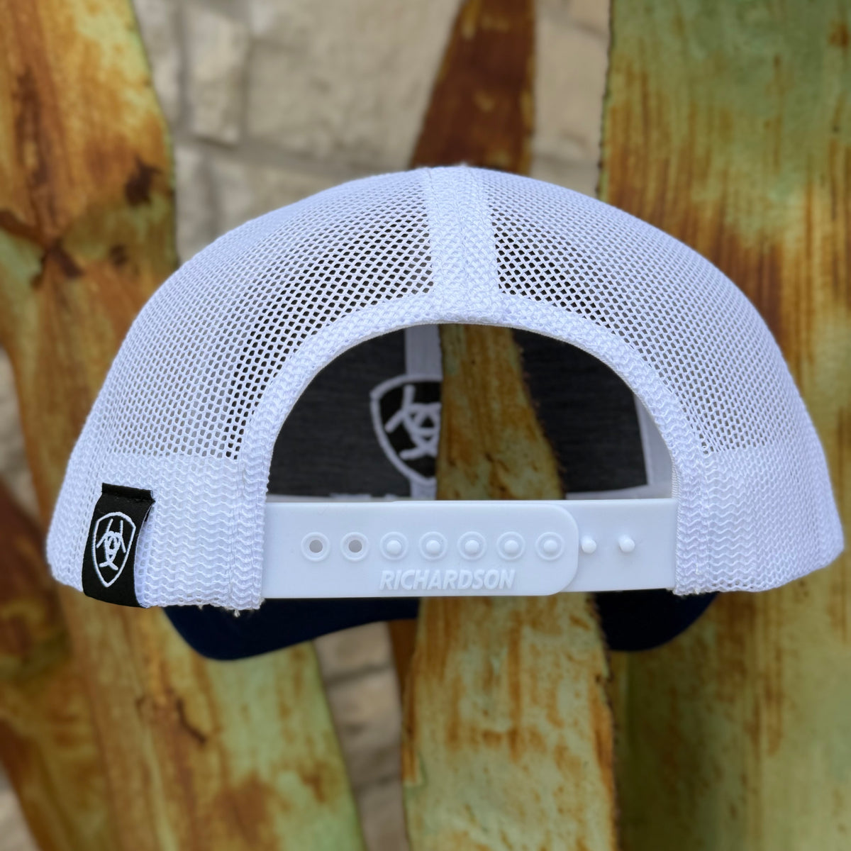 White mesh trucker cap viewed from the back, featuring a white plastic adjustment strap with 'RICHARDSON' printed on it, a black brand patch on the side, and a dark interior lining, set against a rustic wooden background.