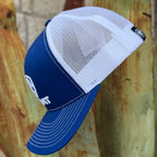 A blue and white trucker-style cap with a curved brim, shown leaning against a rustic wooden surface. The front panel is solid blue with white embroidered lettering, while the back is white mesh.