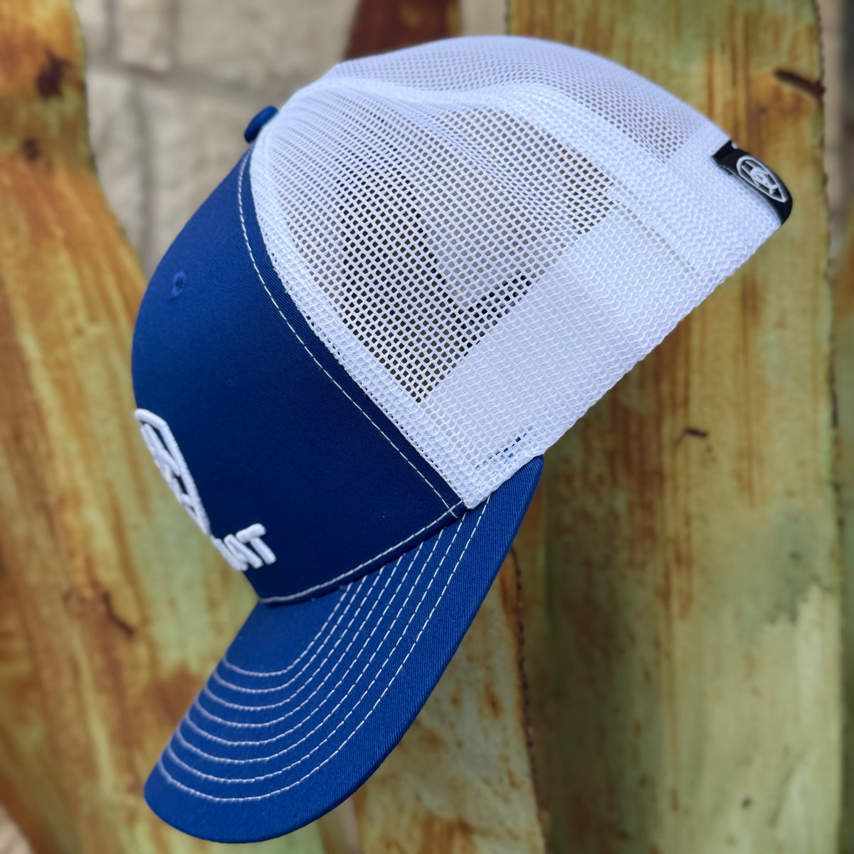 A blue and white trucker-style cap with a curved brim, shown leaning against a rustic wooden surface. The front panel is solid blue with white embroidered lettering, while the back is white mesh.