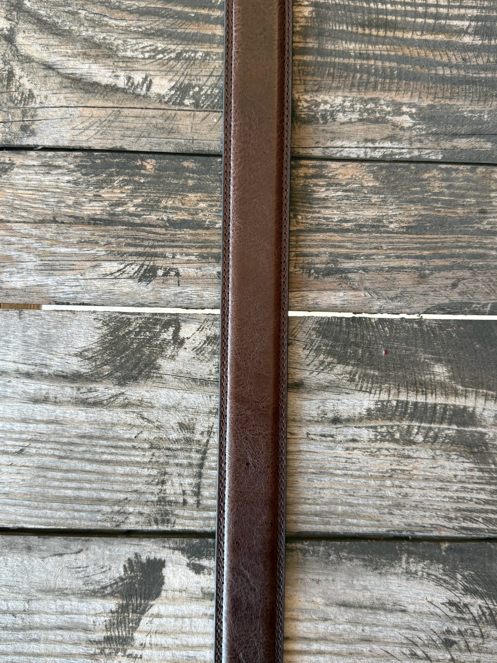A brown leather belt with a silver-toned western-style buckle and decorative tip, coiled on a rustic wooden surface with a distressed red wooden background.