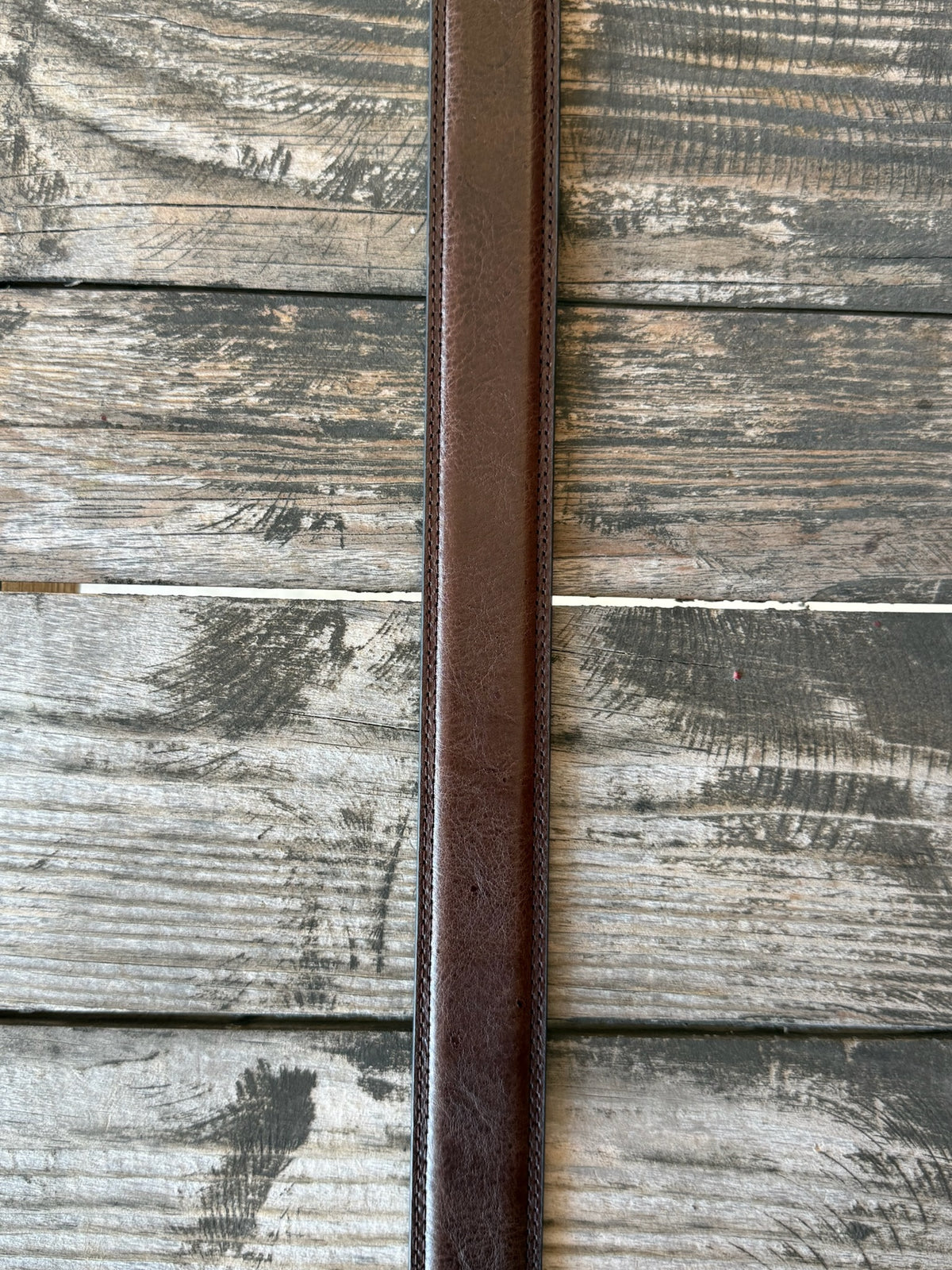 A brown leather belt with a silver-toned western-style buckle and decorative tip, coiled on a rustic wooden surface with a distressed red wooden background.