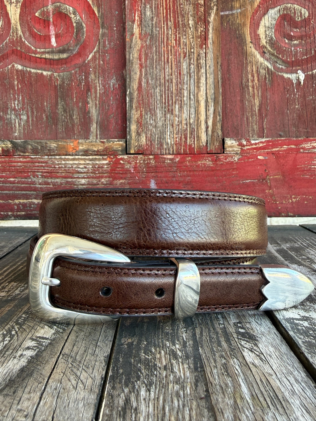A brown leather belt with a silver-toned western-style buckle and decorative tip, coiled on a rustic wooden surface with a distressed red wooden background.