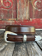 A brown leather belt with a silver-toned western-style buckle and decorative tip, coiled on a rustic wooden surface with a distressed red wooden background.