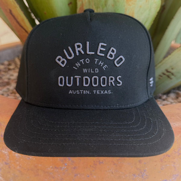 A black structured baseball cap with white embroidered text reading 'BURLEBO INTO THE WILD OUTDOORS AUSTIN, TEXAS.' placed outdoors on a terracotta surface with green plants in the background.