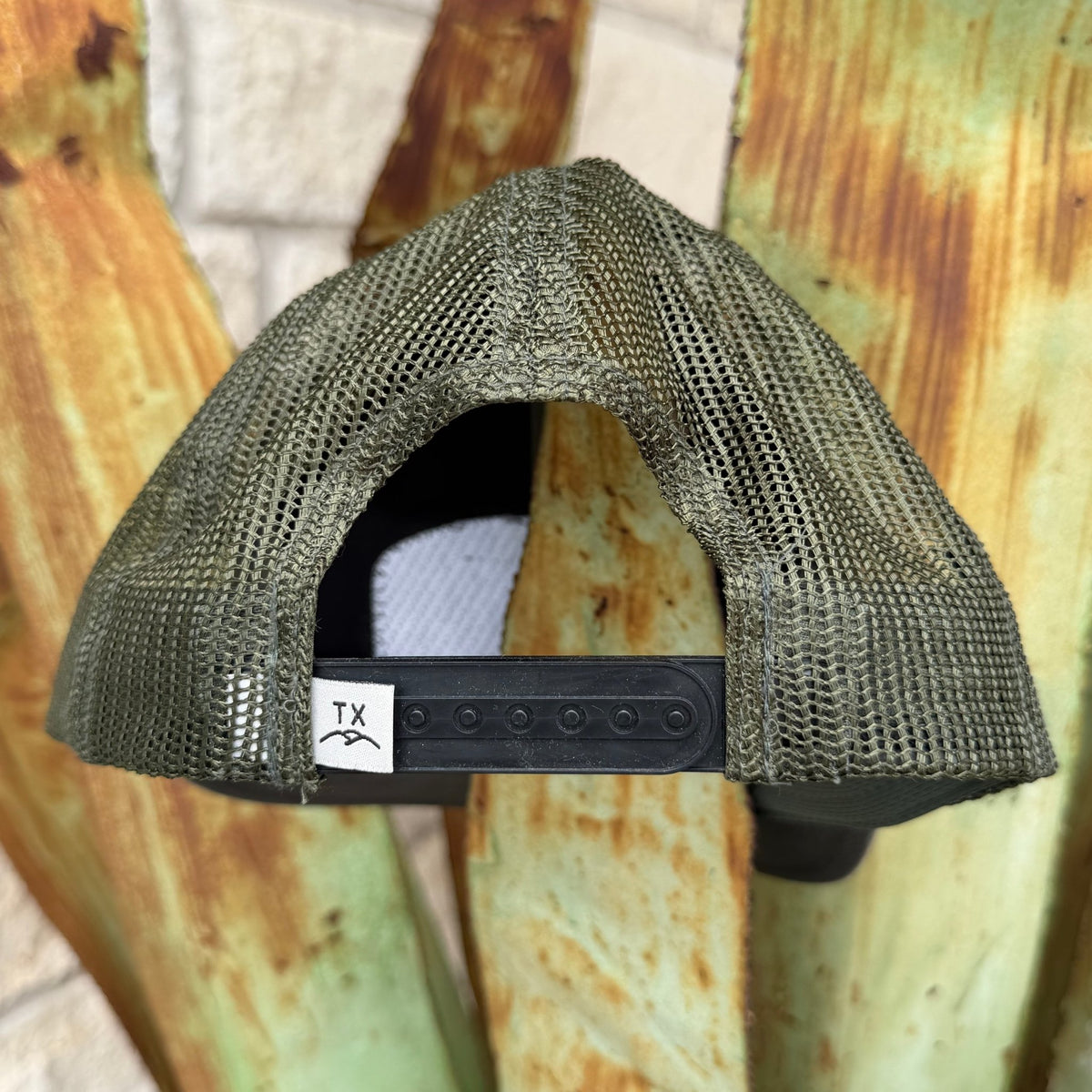 The back view of a trucker-style mesh cap in olive green, featuring a black adjustable strap with a white 'TX' logo patch