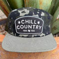 A camouflage trucker hat with a flat brim in olive gray, featuring a front patch that reads 'CHILL COUNTRY AUS TEX' with small palm tree icons and a texas logo. \