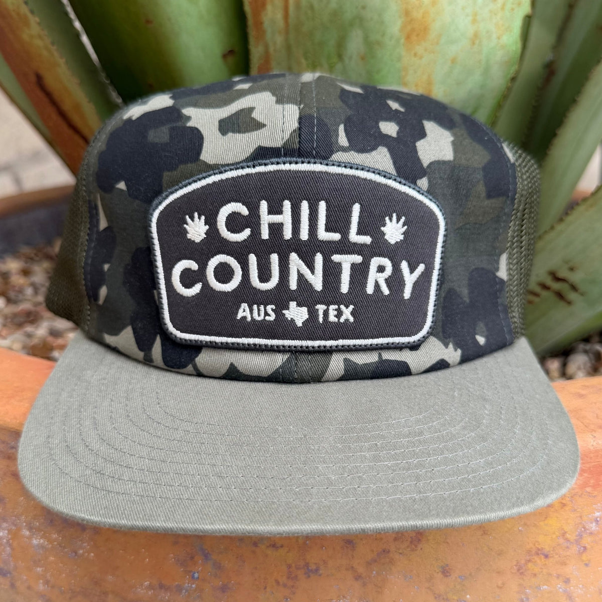A camouflage trucker hat with a flat brim in olive gray, featuring a front patch that reads 'CHILL COUNTRY AUS TEX' with small palm tree icons and a texas logo. \
