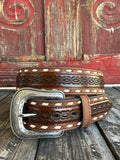 A brown tooled leather belt with white diamond inlays and a large engraved silver buckle, displayed on a rustic wooden surface against a weathered red wooden background.