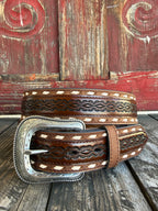 A brown tooled leather belt with white diamond inlays and a large engraved silver buckle, displayed on a rustic wooden surface against a weathered red wooden background.