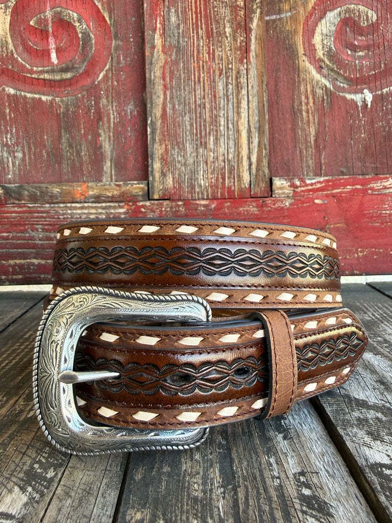 A brown tooled leather belt with white diamond inlays and a large engraved silver buckle, displayed on a rustic wooden surface against a weathered red wooden background.