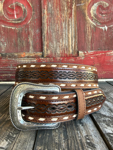 A brown tooled leather belt with white diamond inlays and a large engraved silver buckle, displayed on a rustic wooden surface against a weathered red wooden background.