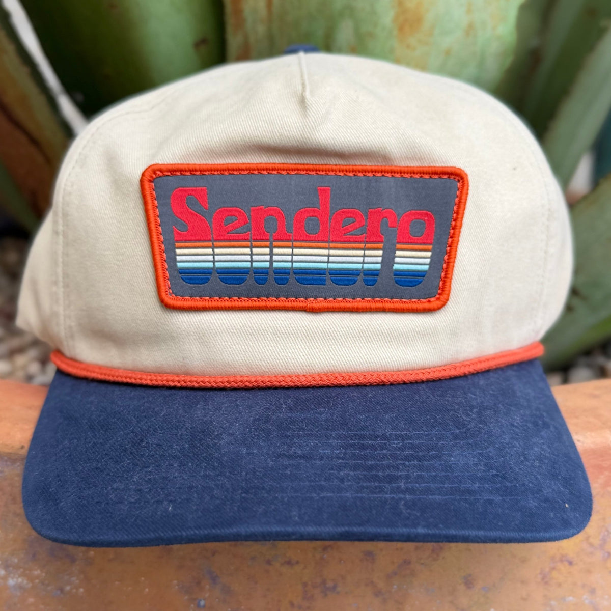 A vintage-style Sendero trucker hat with a beige crown, navy blue visor, and a rectangular embroidered patch on the front featuring the Sendero brand name in red and blue over a striped background, set against a blurred outdoor background.