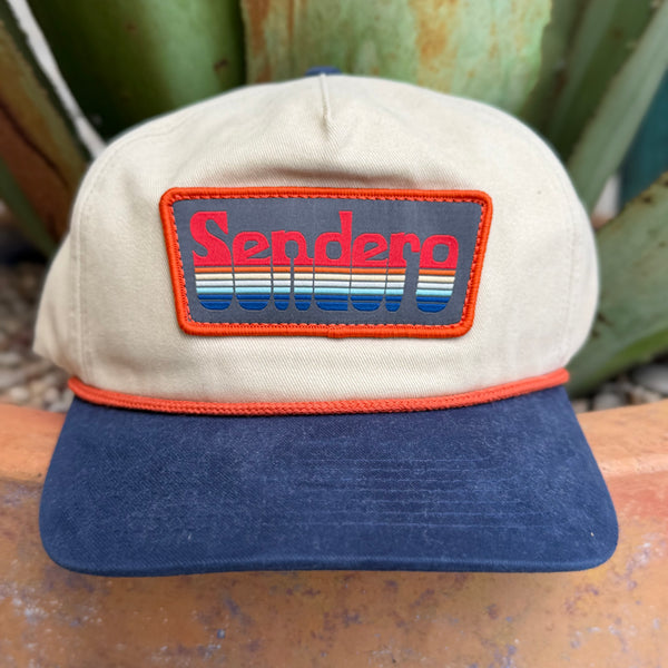 A vintage-style Sendero trucker hat with a beige crown, navy blue visor, and a rectangular embroidered patch on the front featuring the Sendero brand name in red and blue over a striped background, set against a blurred outdoor background.