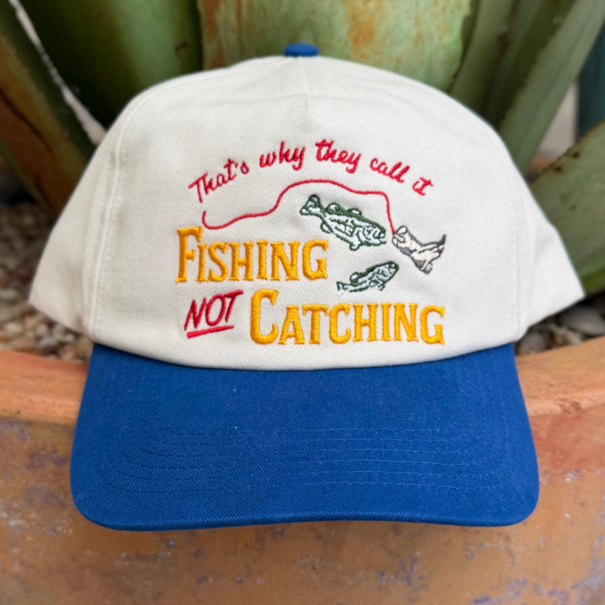 A clean structured five-panel silhouette with a pre-curved blue bill and breathable mesh back. The embroidered fishing graphic and bold lettering run across the front in a natural, blue, and yellow colorway — fun, detailed, and built to hold up wear after wear. Snapback closure at the back for a dialed-in fit.