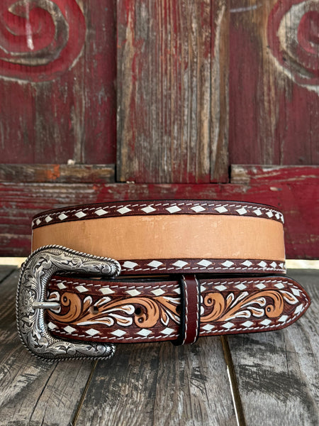 A western-style leather belt with a silver engraved buckle, featuring a tan center stripe, dark brown edges, and hand-tooled floral scrollwork in tan and white, resting on a rustic wooden surface.