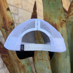 A white trucker-style cap viewed from the back, featuring a white mesh back and an adjustable white plastic snap strap. A small black brand tag is visible on the left side of the strap.
