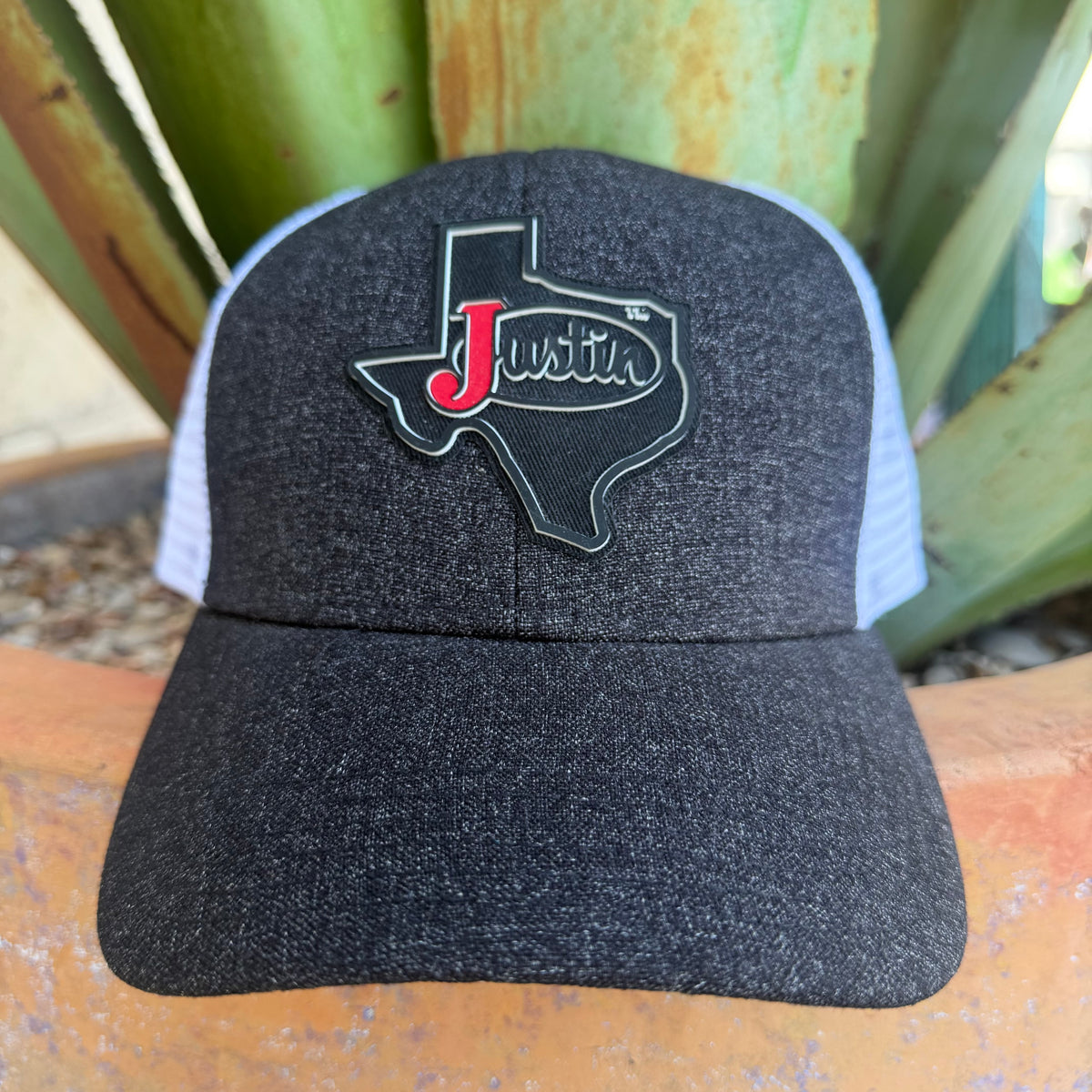 A dark heather gray trucker hat with a Texas-shaped patch on the front. The patch is black with a red script 'J' and the word 'Justin'