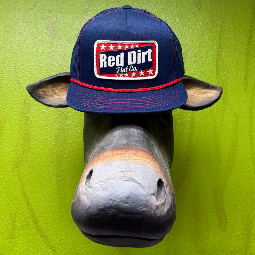 Rep the brand that knows Western. This Red Dirt Hat Co. trucker cap keeps it classic with a structured navy front, breathable mesh back, and a bold embroidered patch that does all the talking. The red rope accent cord along the brim adds just enough detail to set it apart from every other cap on the rack. One size, no fuss, all attitude.