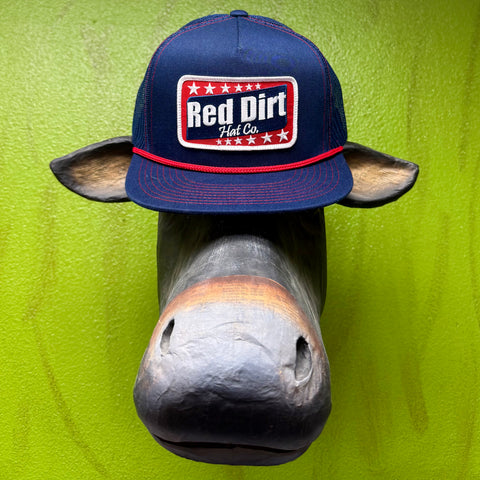 Rep the brand that knows Western. This Red Dirt Hat Co. trucker cap keeps it classic with a structured navy front, breathable mesh back, and a bold embroidered patch that does all the talking. The red rope accent cord along the brim adds just enough detail to set it apart from every other cap on the rack. One size, no fuss, all attitude.