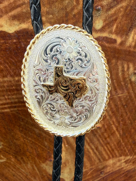 Men’s Montana Silversmiths silver and gold oval Texas bolo tie, featuring engraved floral scrolls, gold rope edge, and black braided leather cord with silver arrow tips – BT22. Available at Blair's Western Wear, Marble Falls, TX.