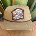 A khaki beige flat-brim cap with a brown embroidered patch on the front that reads 'PANCHO PALOMAS' and shows two doves in sombreros and rifles 