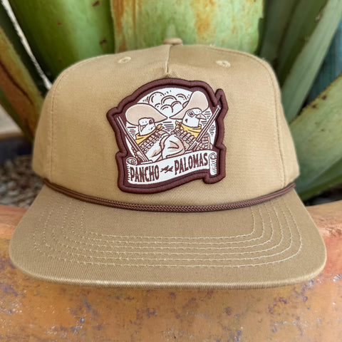A khaki beige flat-brim cap with a brown embroidered patch on the front that reads 'PANCHO PALOMAS' and shows two doves in sombreros and rifles 