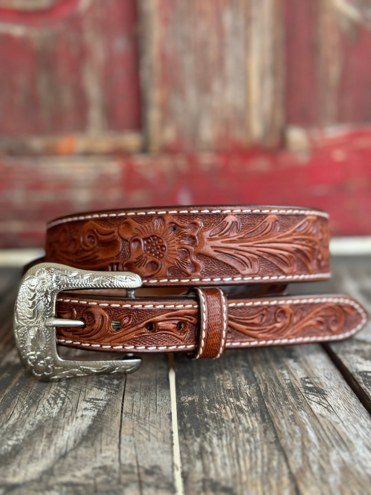 A brown tooled leather belt with floral embossing and a silver western-style buckle, displayed on a rustic wooden surface with a blurred red wooden background.