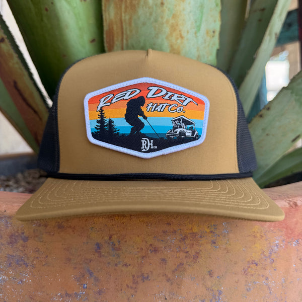 A tan and black trucker hat with a 'Red Dirt Hat Co.' patch on the front, 