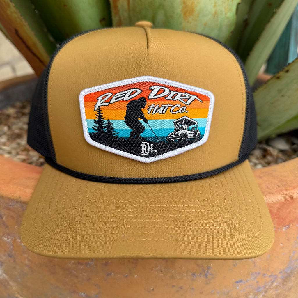 A tan and black mesh trucker hat with a 'Red Dirt Hat Co.' embroidered patch on the front, displayed outdoors on a terracotta surface with green succulents in the background.