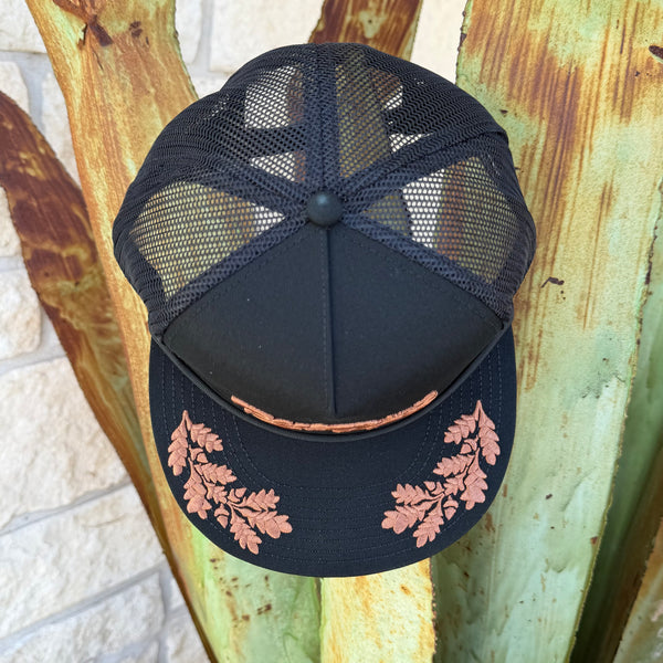 A black trucker-style cap with a black mesh back and a black brim featuring two copper leaf designs on the front and back,