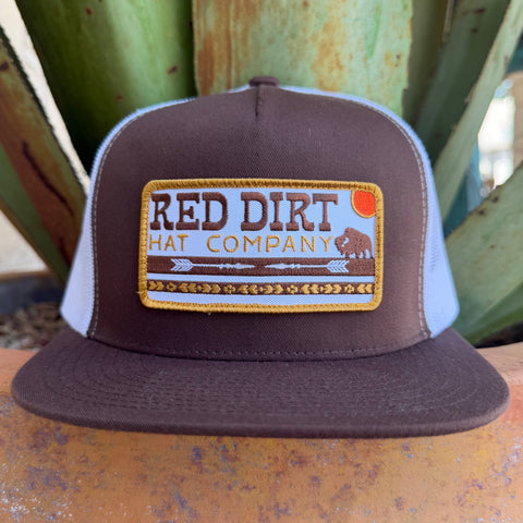 A brown and white trucker hat with a rectangular embroidered patch reading 'RED DIRT HAT COMPANY' on the front, 