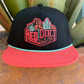 A black trucker hat with a coral-pink flat brim and a mint green edge, featuring a red and pink embroidered 'Red Dirt Hat Co' logo with desert illustrations on the front. 