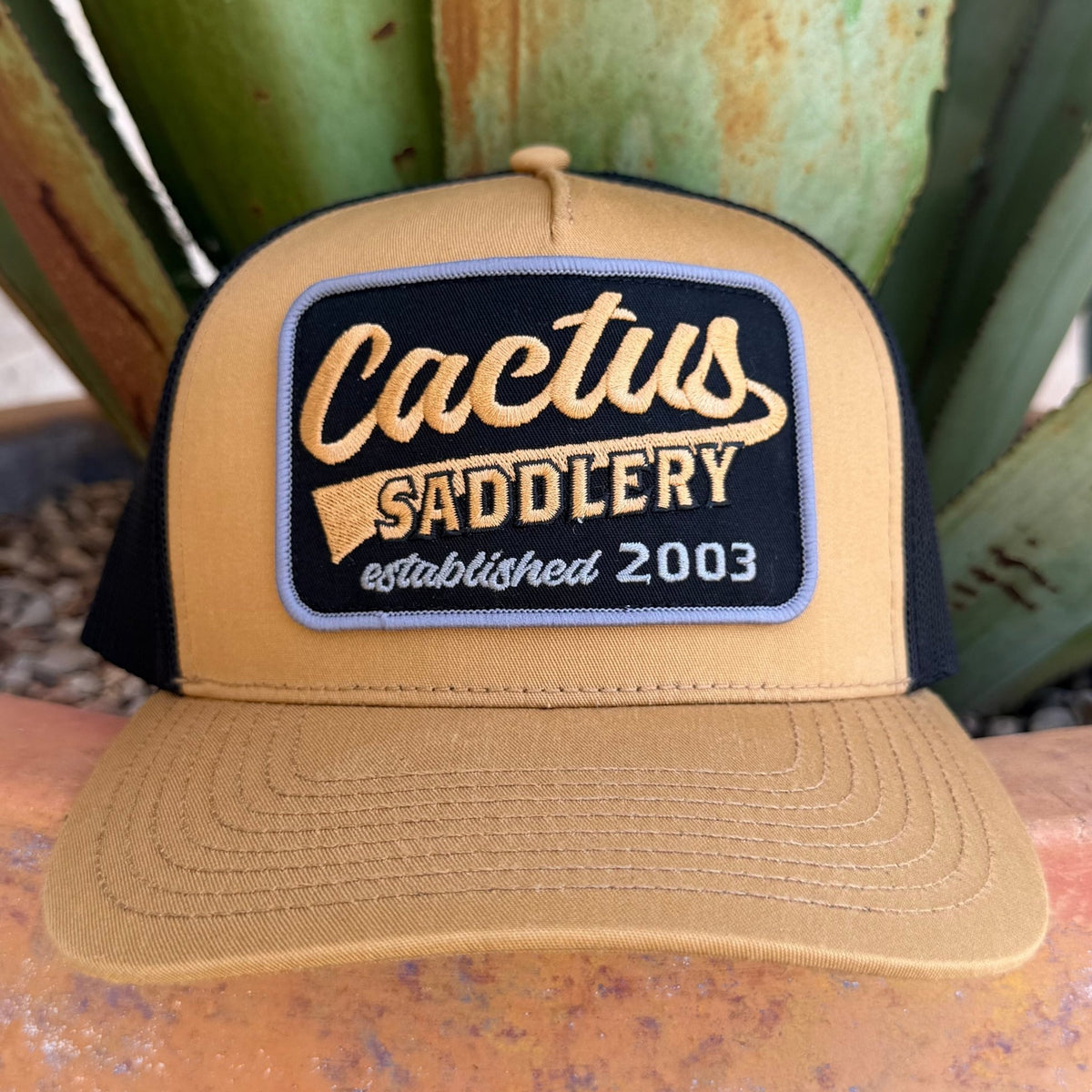 A tan and black trucker hat with a front patch reading 'Cactus Saddlery established 2003"