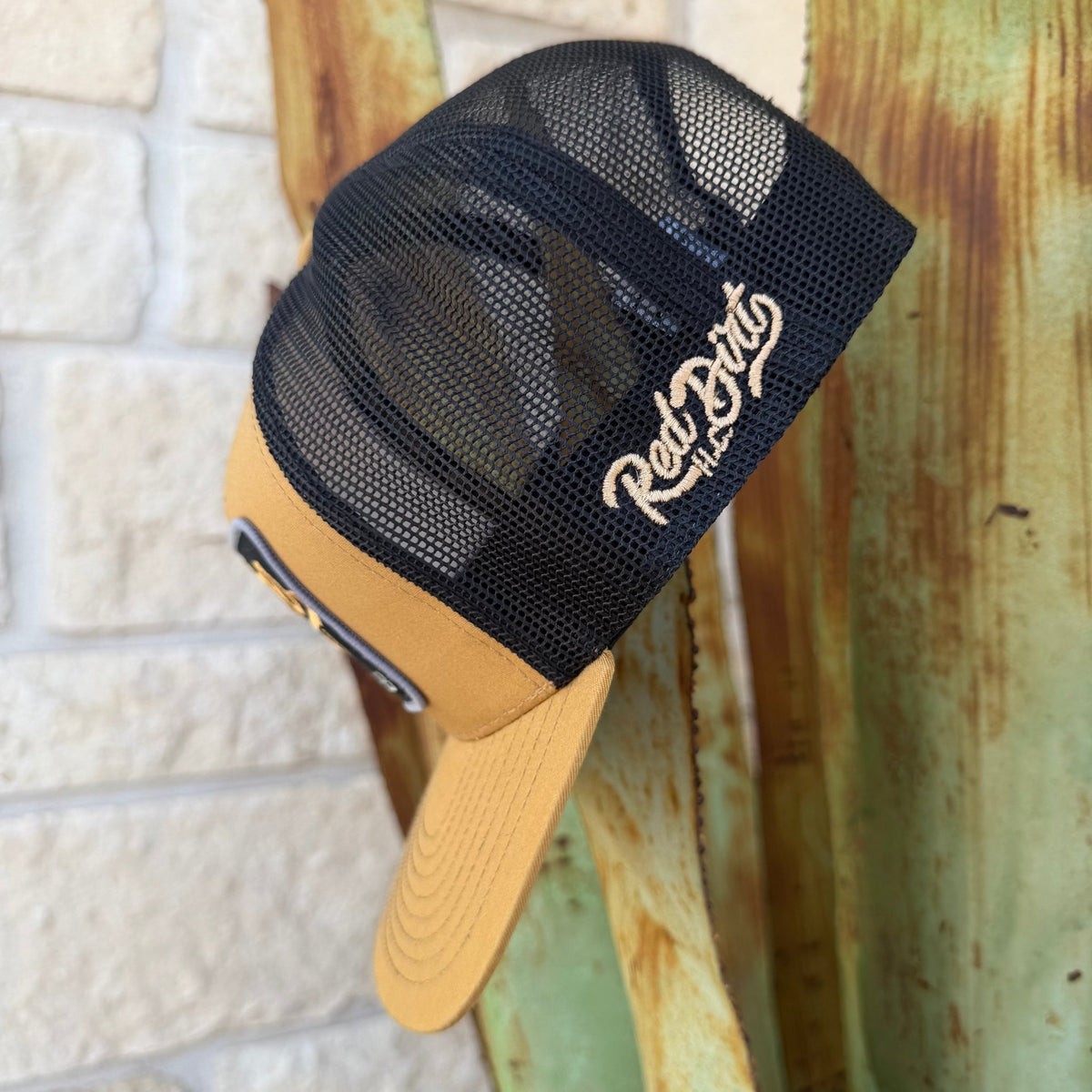 A trucker-style cap with a black mesh back and a tan visor, featuring a embroidered 'Red Dirt' logo on the side