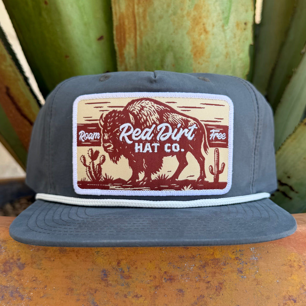 A dark gray snapback cap featuring a rectangular embroidered front patch with a bison, cacti, and the text 'Red Dirt Hat Co.' and 'Roam Free.