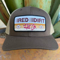 A brown trucker hat with a beige mesh back, featuring a rectangular front patch that reads 'RED DIRT Hat Co.' in red and black lettering with an aztec inspired pattern in golden colors 