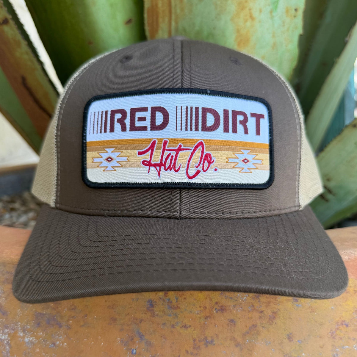 A brown trucker hat with a beige mesh back, featuring a rectangular front patch that reads 'RED DIRT Hat Co.' in red and black lettering with an aztec inspired pattern in golden colors 