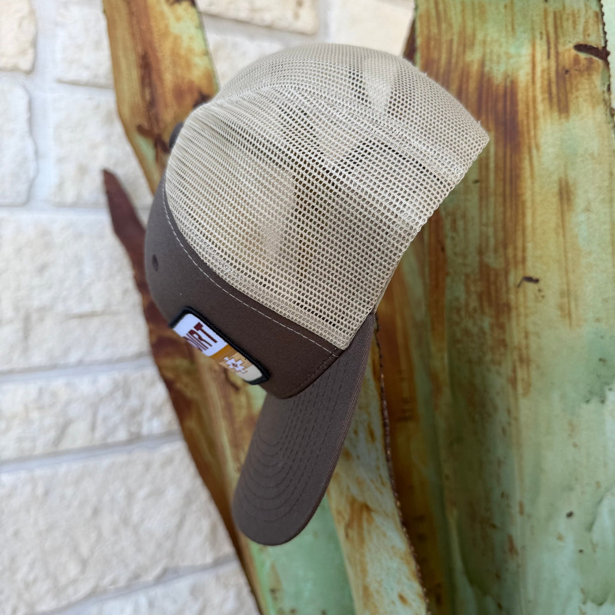 A brown trucker hat with a beige mesh back, featuring a rectangular front patch that reads 'RED DIRT Hat Co.' in red and black lettering with an aztec inspired pattern in golden colors 