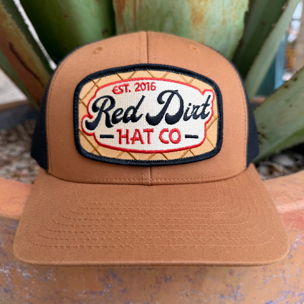 A tan trucker-style cap with black mesh side panels and a large embroidered front patch reading 'Red Dirt HAT CO' and 'EST. 2016'.