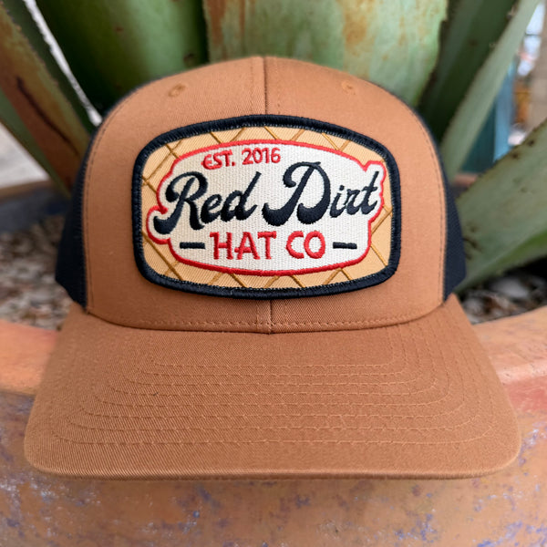 A tan trucker-style cap with black mesh side panels and a large embroidered front patch reading 'Red Dirt HAT CO' and 'EST. 2016'.