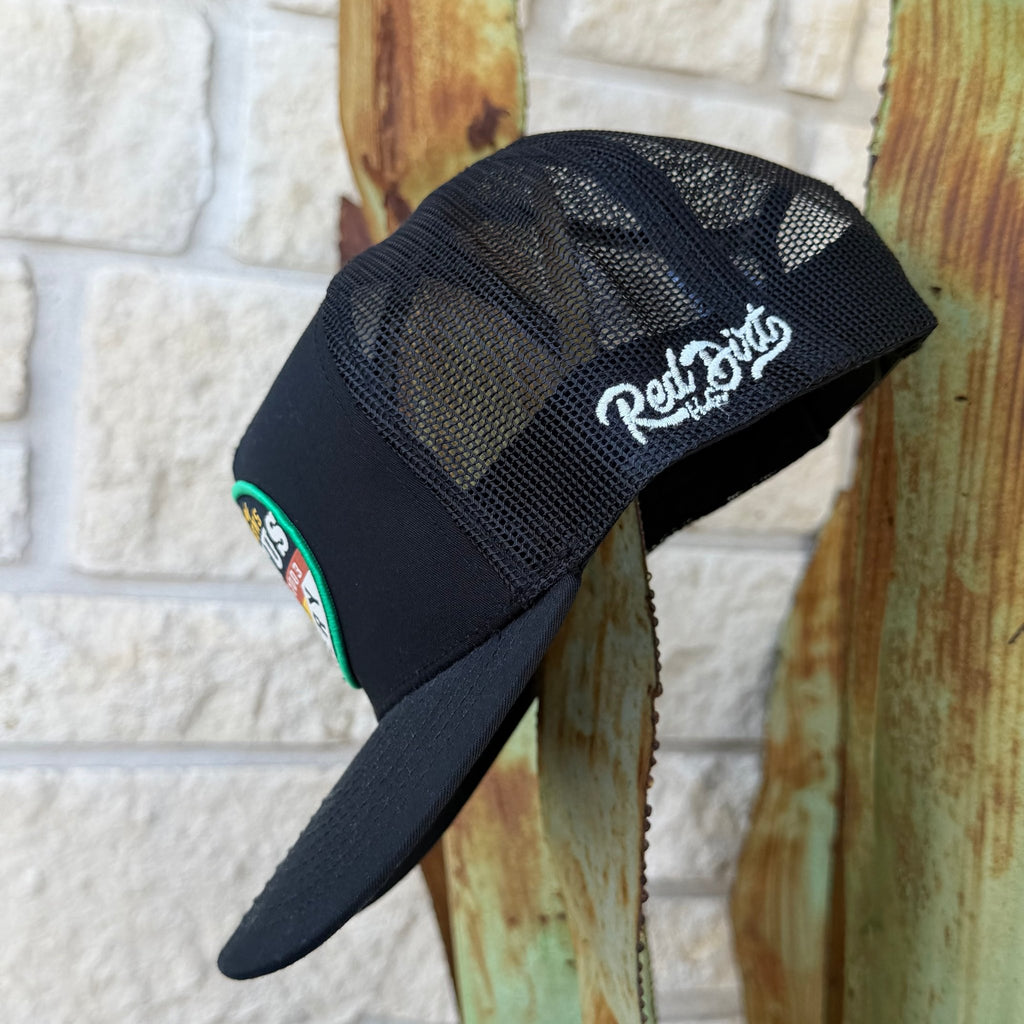 A black trucker-style cap with a mesh back and a curved brim, leaning against a rustic wooden post in front of a light stone wall. The brim has a white script logo reading 'Red Dirt' with smaller text below, and the side panel shows a small multicolor patch.