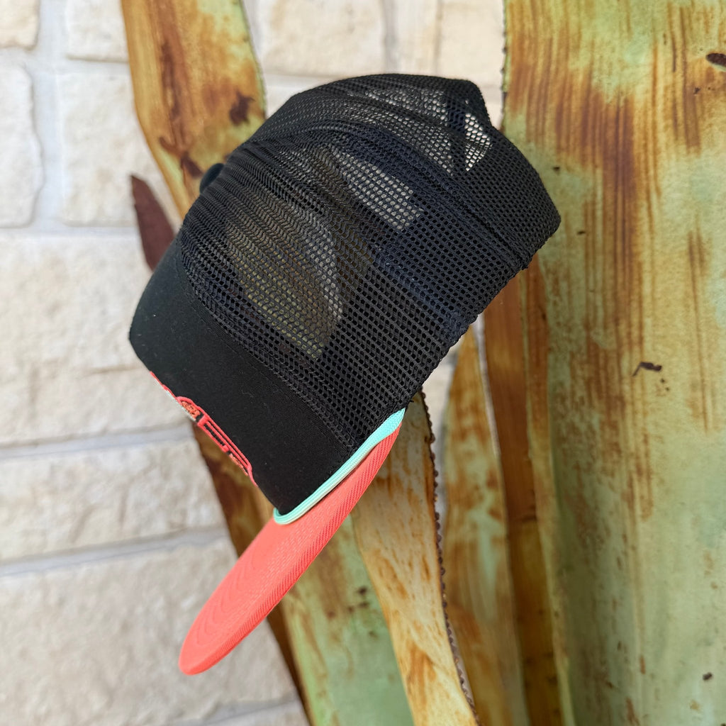 A black trucker hat with a coral-pink flat brim and a mint green edge, featuring a red and pink embroidered 'Red Dirt Hat Co' logo with desert illustrations on the front. 