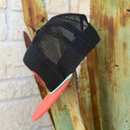 A black trucker hat with a coral-pink flat brim and a mint green edge, featuring a red and pink embroidered 'Red Dirt Hat Co' logo with desert illustrations on the front. 