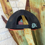 A black trucker hat with a coral-pink flat brim and a mint green edge, featuring a red and pink embroidered 'Red Dirt Hat Co' logo with desert illustrations on the front. 