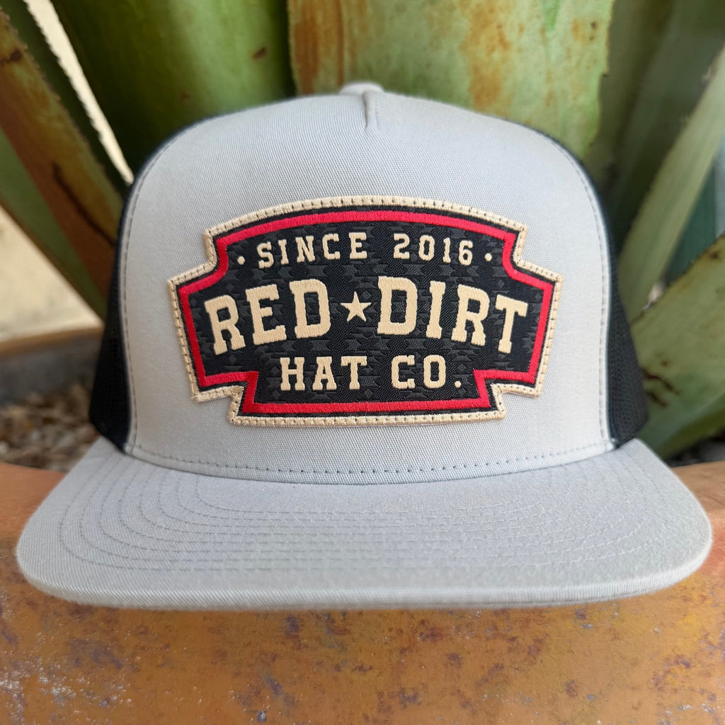 A light gray and black trucker hat with a red and black embroidered front patch reading 'SINCE 2016 RED DIRT HAT CO.' in off white lettering
