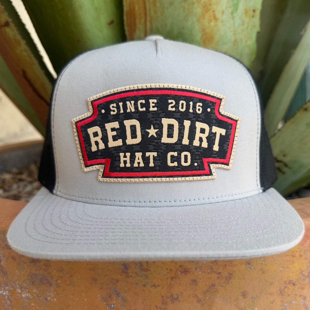 A light gray and black trucker hat with a red and black embroidered front patch reading 'SINCE 2016 RED DIRT HAT CO.' in off white lettering