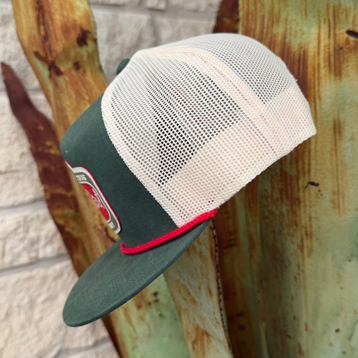 A dark green and off-white mesh trucker hat with a red accent stripe and a small embroidered patch on the brim