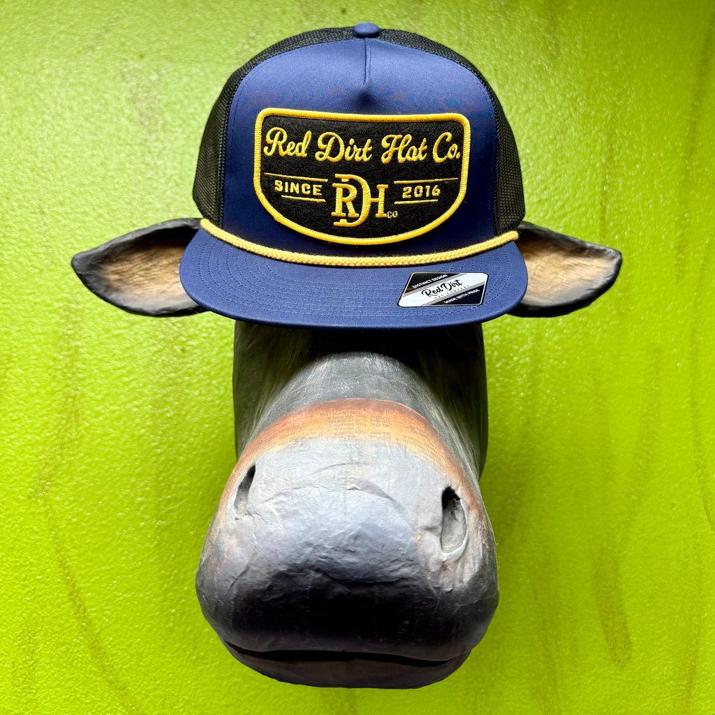 Men’s Red Dirt Hat Co. mesh trucker cap in navy and black, featuring an embroidered yellow logo patch and flat bill with gold rope detail – RDHC441. Available at Blair’s Western Wear, Marble Falls, TX.