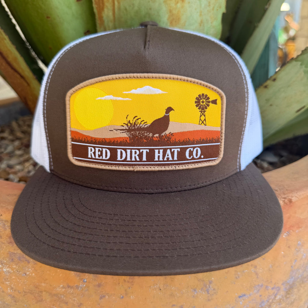 A brown and white trucker hat with a front patch reading 'RED DIRT HAT CO.' and featuring a sunset landscape with a turkey and windmill, 