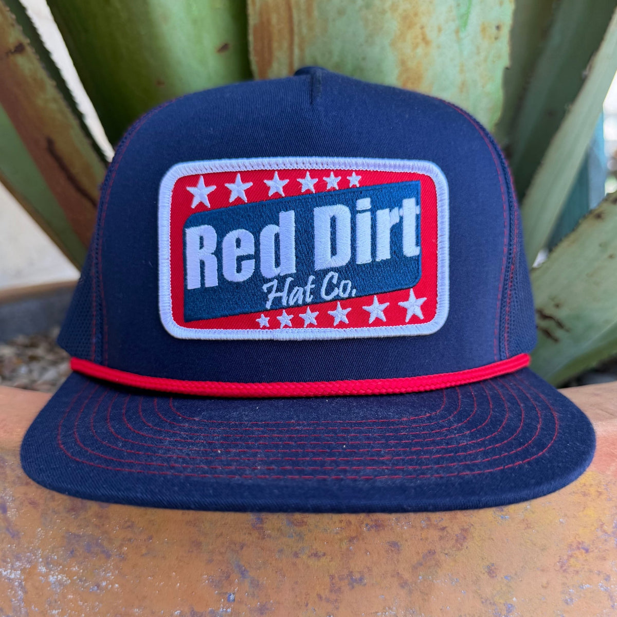 This Red Dirt Hat Co. trucker keeps it classic with a structured navy front, breathable mesh back, and a bold embroidered patch that does all the talking. The red rope accent cord along the brim adds just enough detail to set it apart from every other cap on the rack.